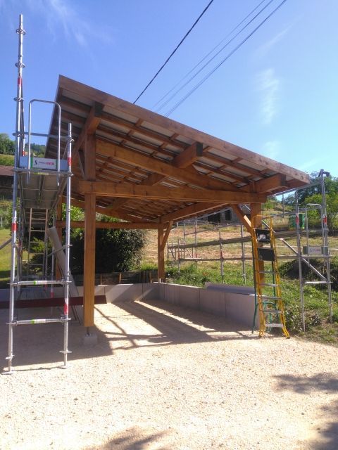 Abri en bois ouvert sur les côtés, avec un toit en pente et un échafaudage métallique, dans un espace extérieur ensoleillé.