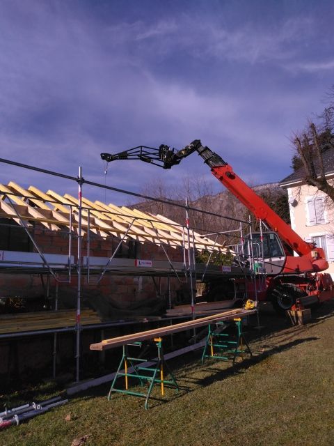 Une grue rouge soulève une longue poutre à côté d'un bâtiment en construction partiellement couvert.