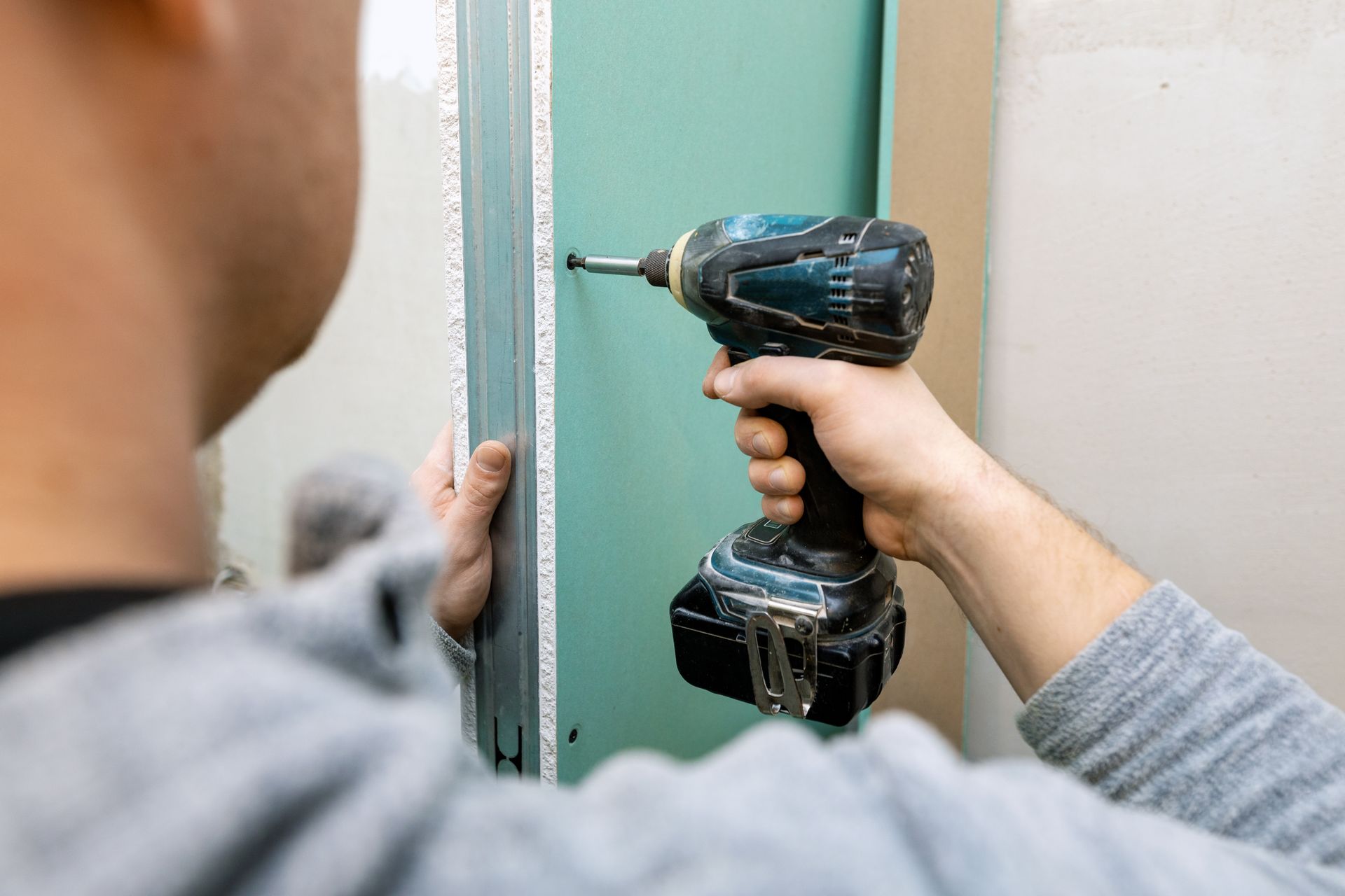 Une personne utilise une perceuse sans fil pour visser une règle métallique sur un mur lors de travaux de rénovation de sa maison.