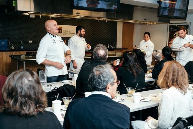 Un grupo de personas están sentadas en mesas en una cocina viendo una presentación de chefs.