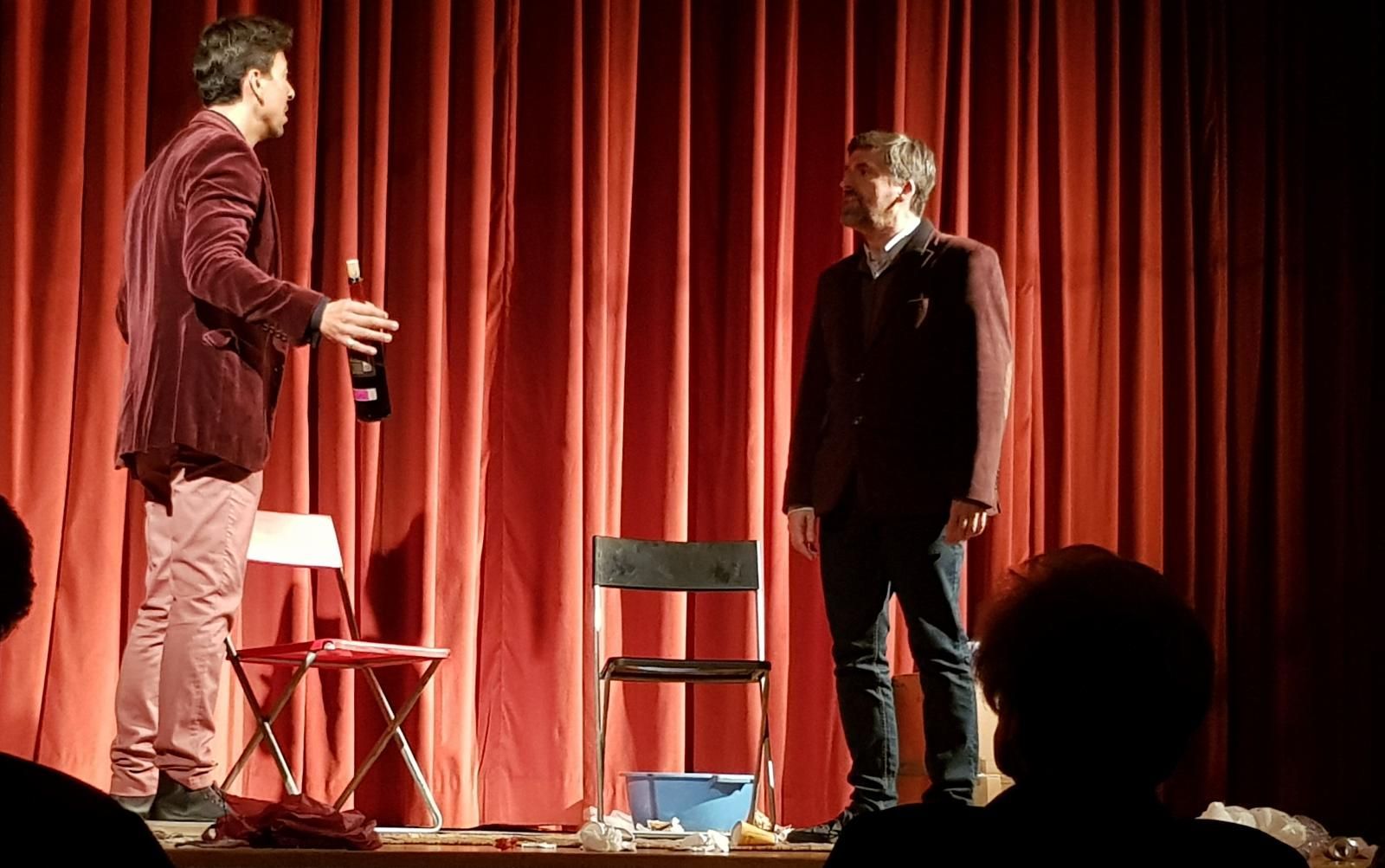 Dos hombres están de pie en un escenario frente a una cortina roja.