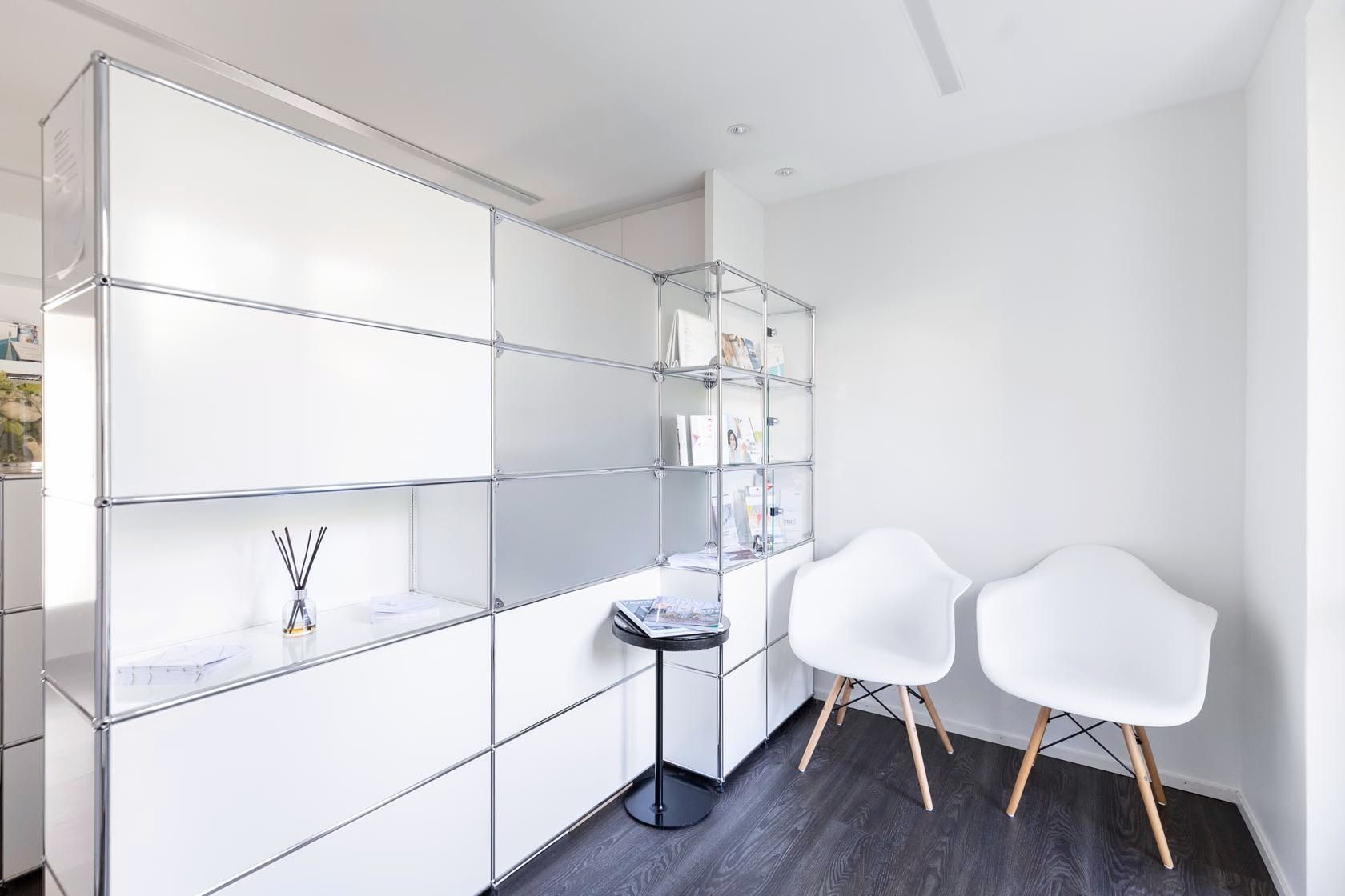 Ein Wartezimmer mit zwei weißen Stühlen und einem Regal.