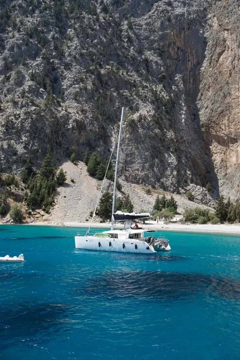 Catamarán navegando en aguas turquesas cerca de una playa con acantilado rocoso.