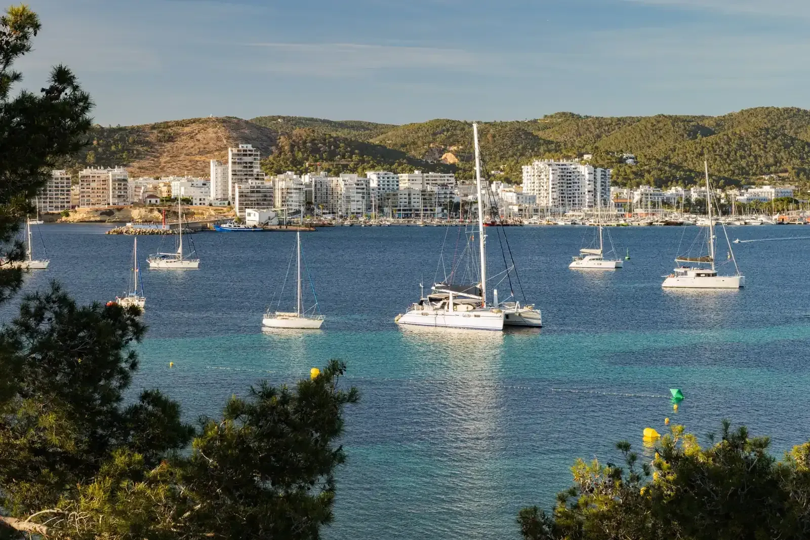 Barcos en tranquilas aguas turquesas, edificios blancos y colinas verdes en un pueblo costero.