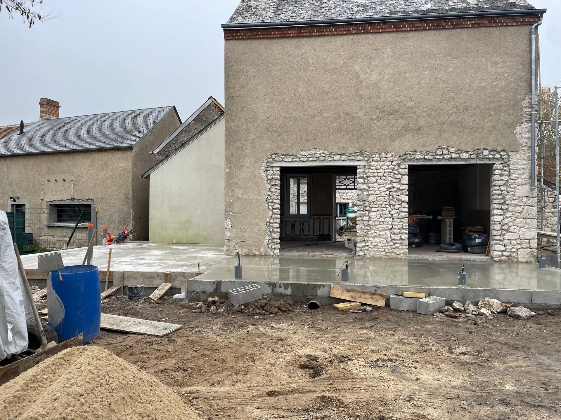 Extérieur d'une maison de campagne avec cour non aménagée, baril bleu et matériaux de construction devant.