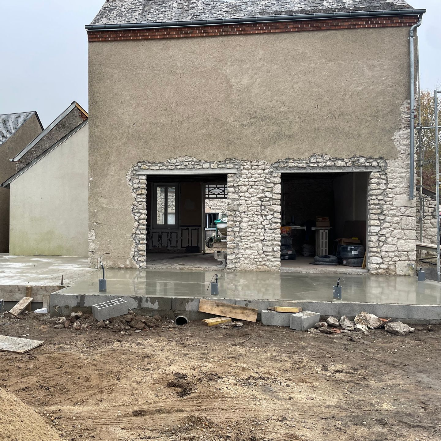 Maison en pierre partiellement rénovée avec deux grandes ouvertures pour des baies vitrées et chantier boueux devant.