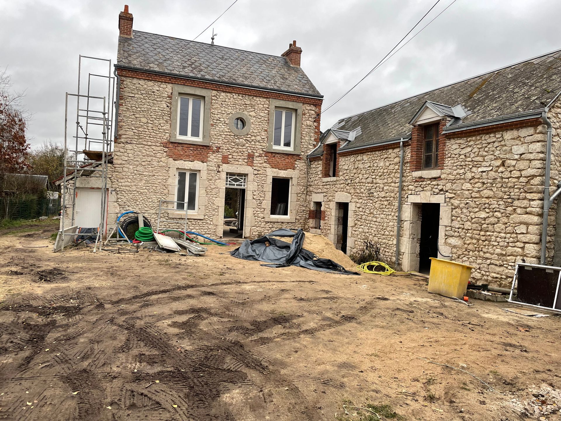 Rénovation d'une maison rurale en pierre avec échafaudages, cour boueuse et débris de construction.