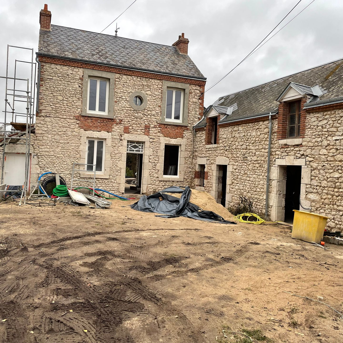 Maison en pierre en cours de rénovation : échafaudages, débris et cour en terre.