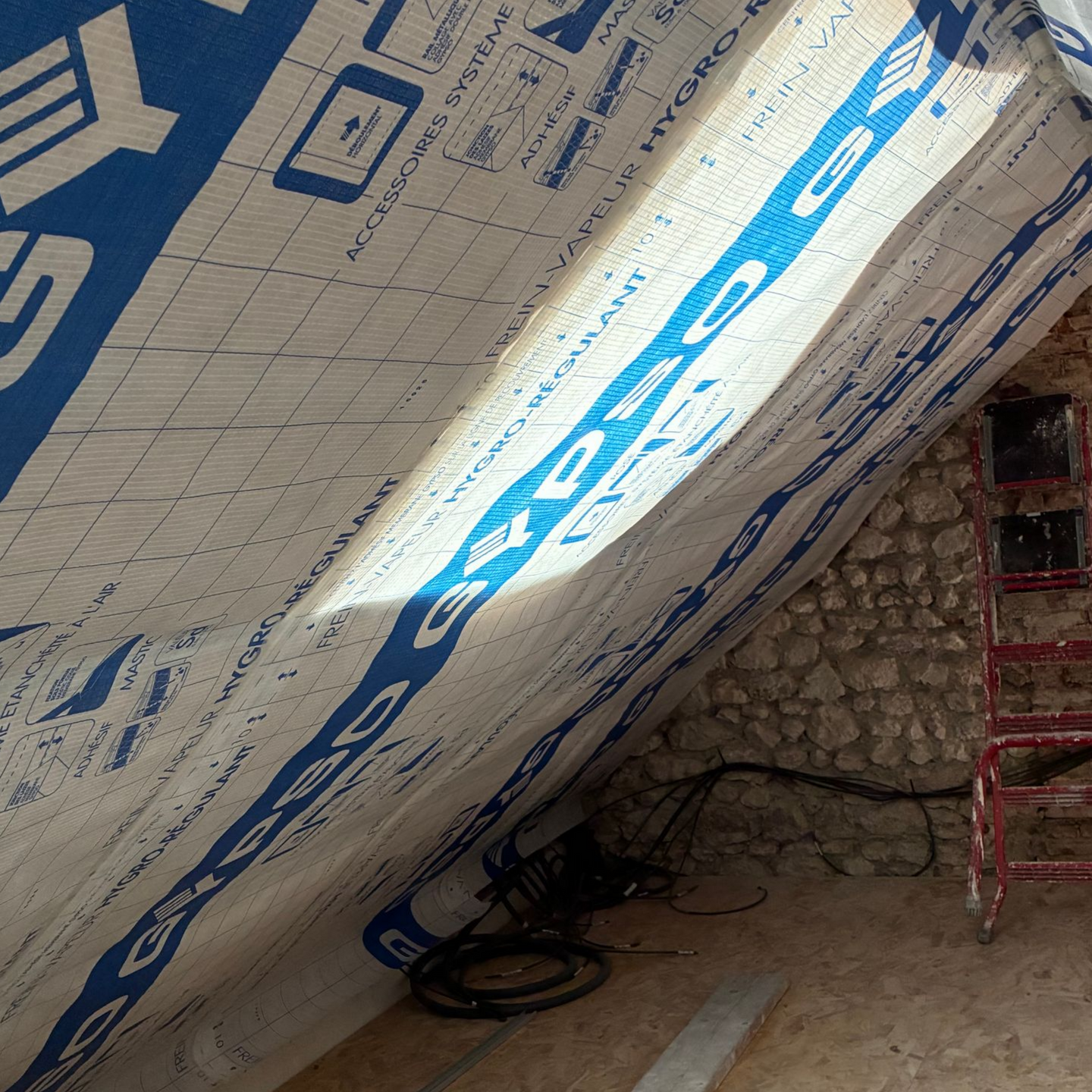 Grenier partiellement isolé avec des panneaux de mousse, fils électriques apparents et une échelle rouge appuyée contre le mur.