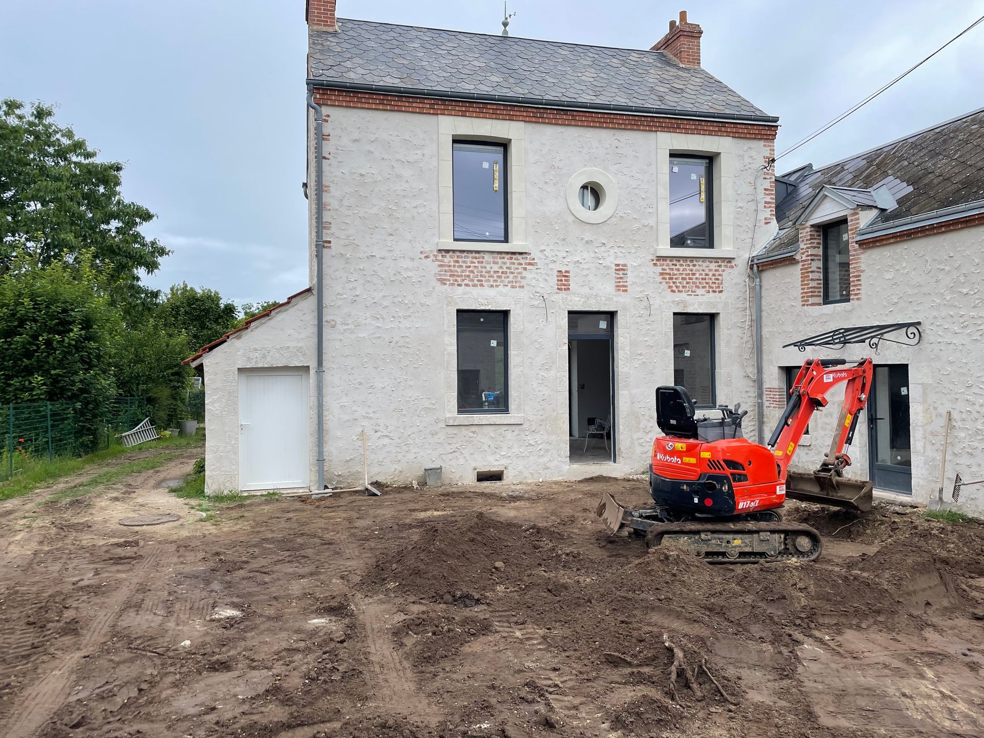 Maison partiellement rénovée avec cour boueuse et petite pelleteuse devant.