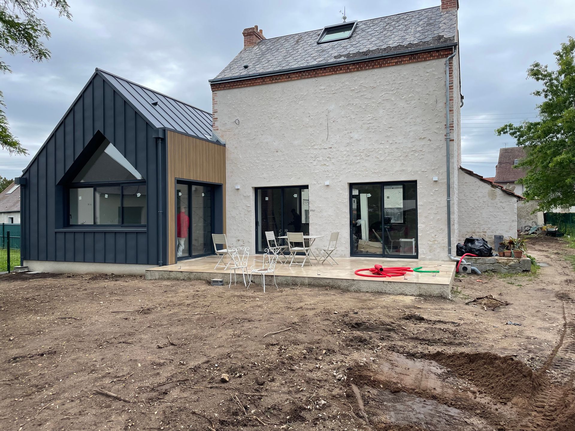 Maison ancienne rénovée avec une extension revêtue de noir et des chaises sur une terrasse.