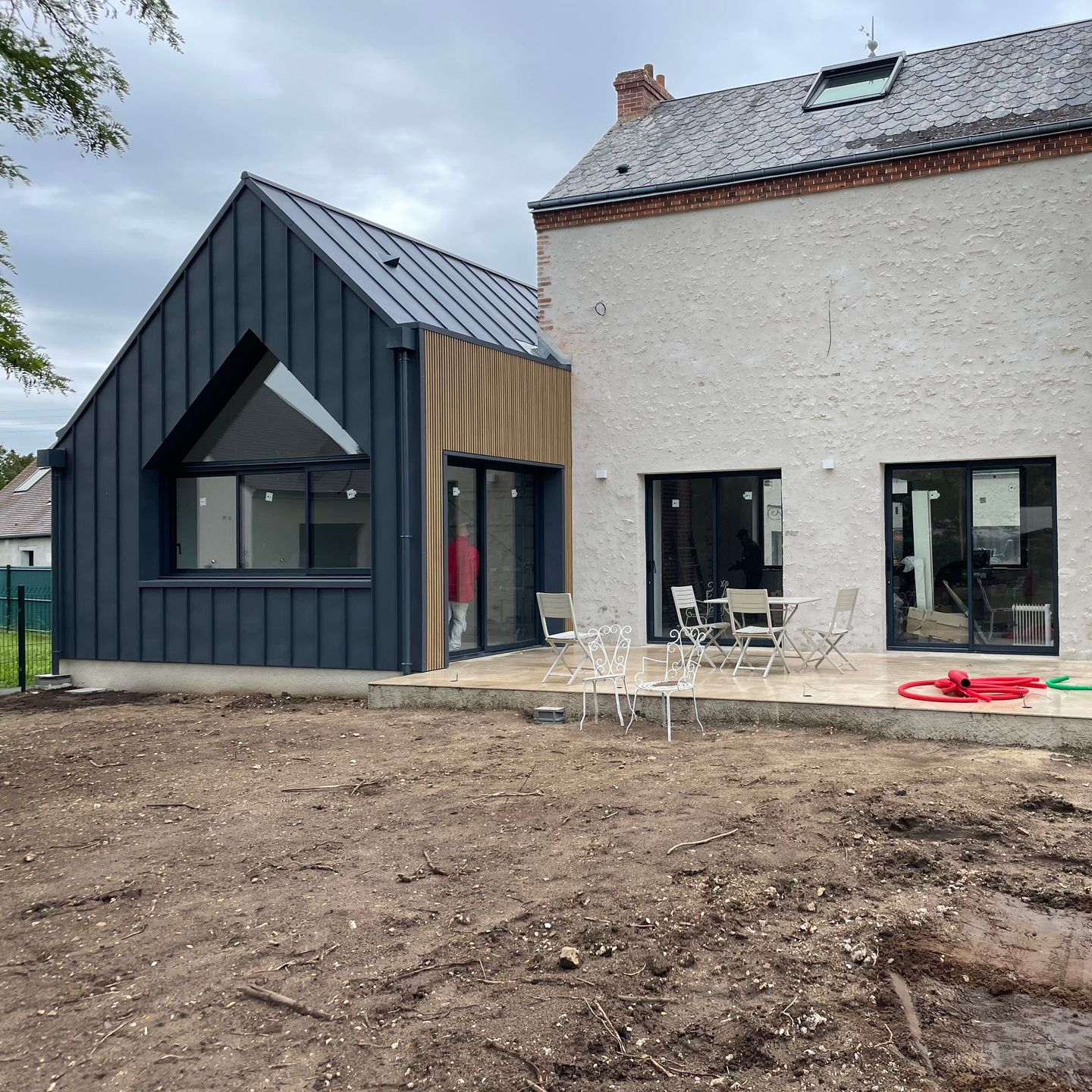 Extérieur de maison moderne avec extension à bardage sombre, mur en pierre claire et cour nue.