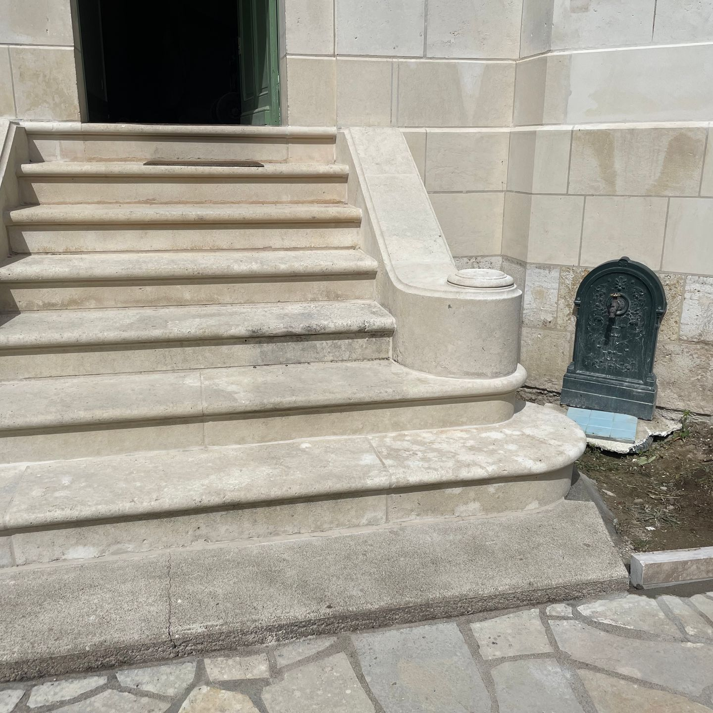 Des marches en pierre mènent à l'entrée d'un bâtiment, avec une rampe incurvée et une petite fontaine murale de jardin sur le côté.