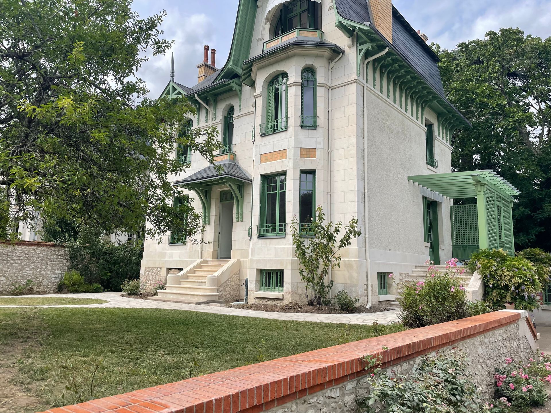 Maison blanche de style victorien avec des boiseries vertes, un perron et un mur de briques rouges dans une cour arborée.