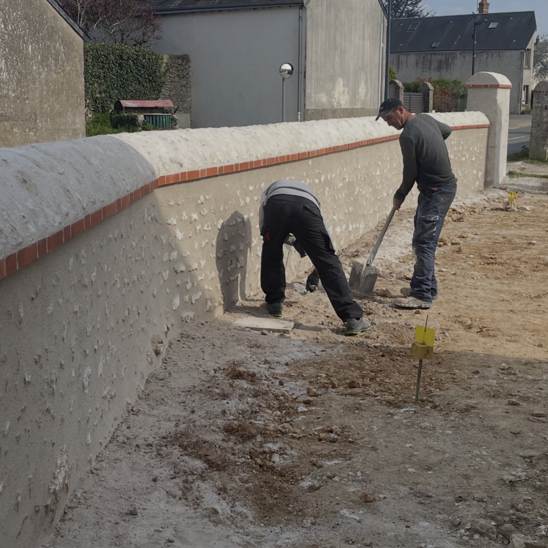 Deux ouvriers travaillent à côté d'un mur de pierre rénové dans une cour en terre battue.