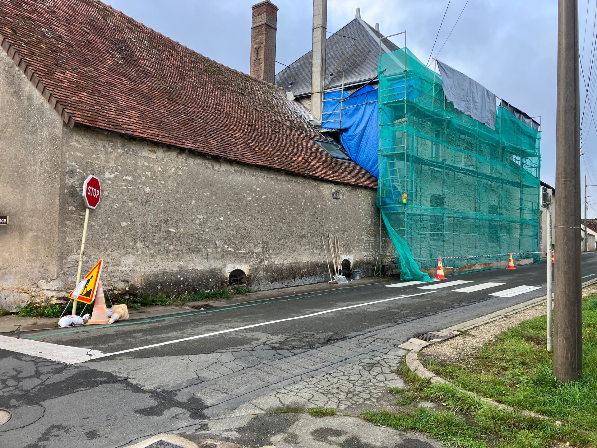 Un vieux bâtiment en pierre en cours de restauration, protégé par des filets verts et des bâches bleues, borde une rue étroite.