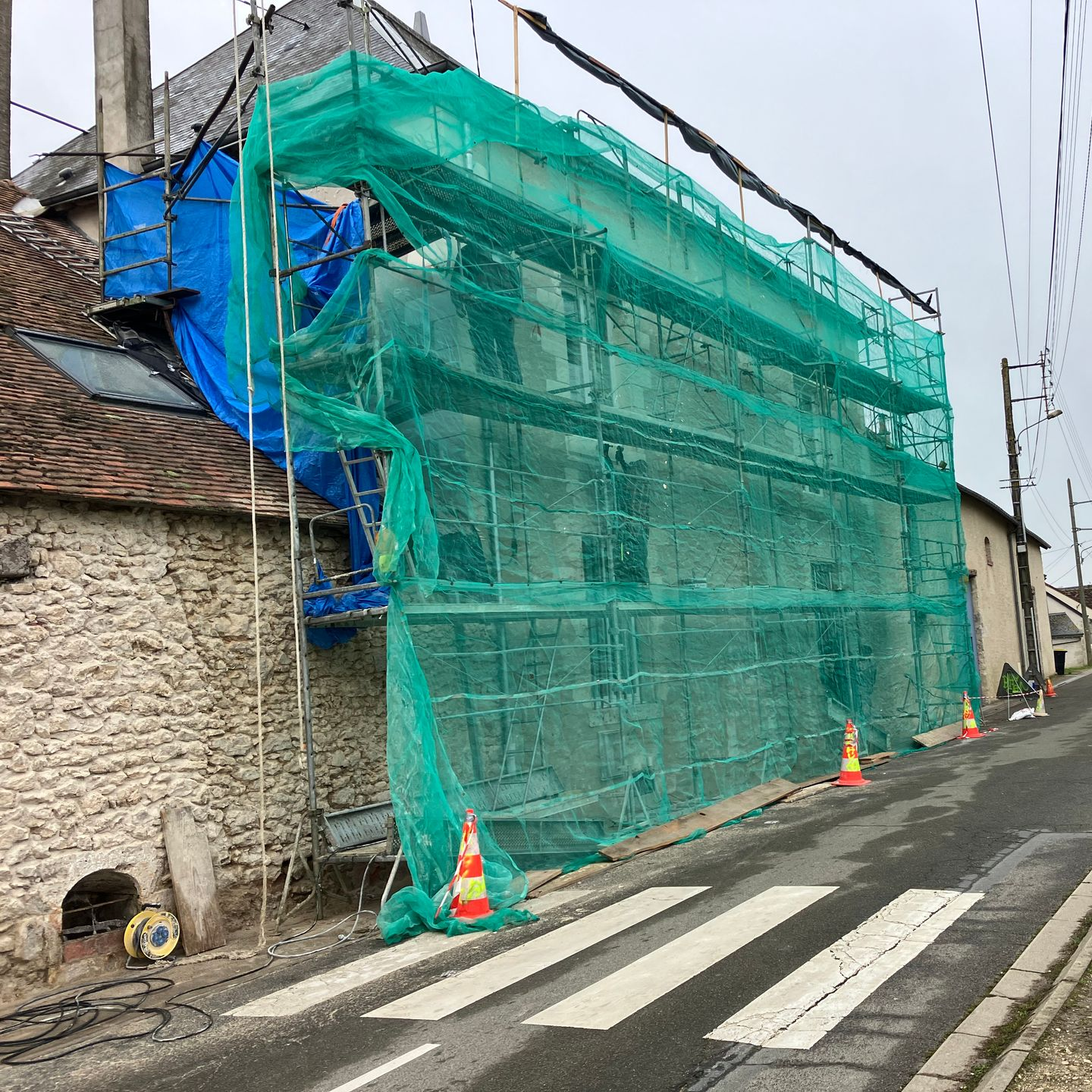 Bâtiment en échafaudage recouvert de filets de sécurité verts, bordant une rue étroite avec un passage piéton.