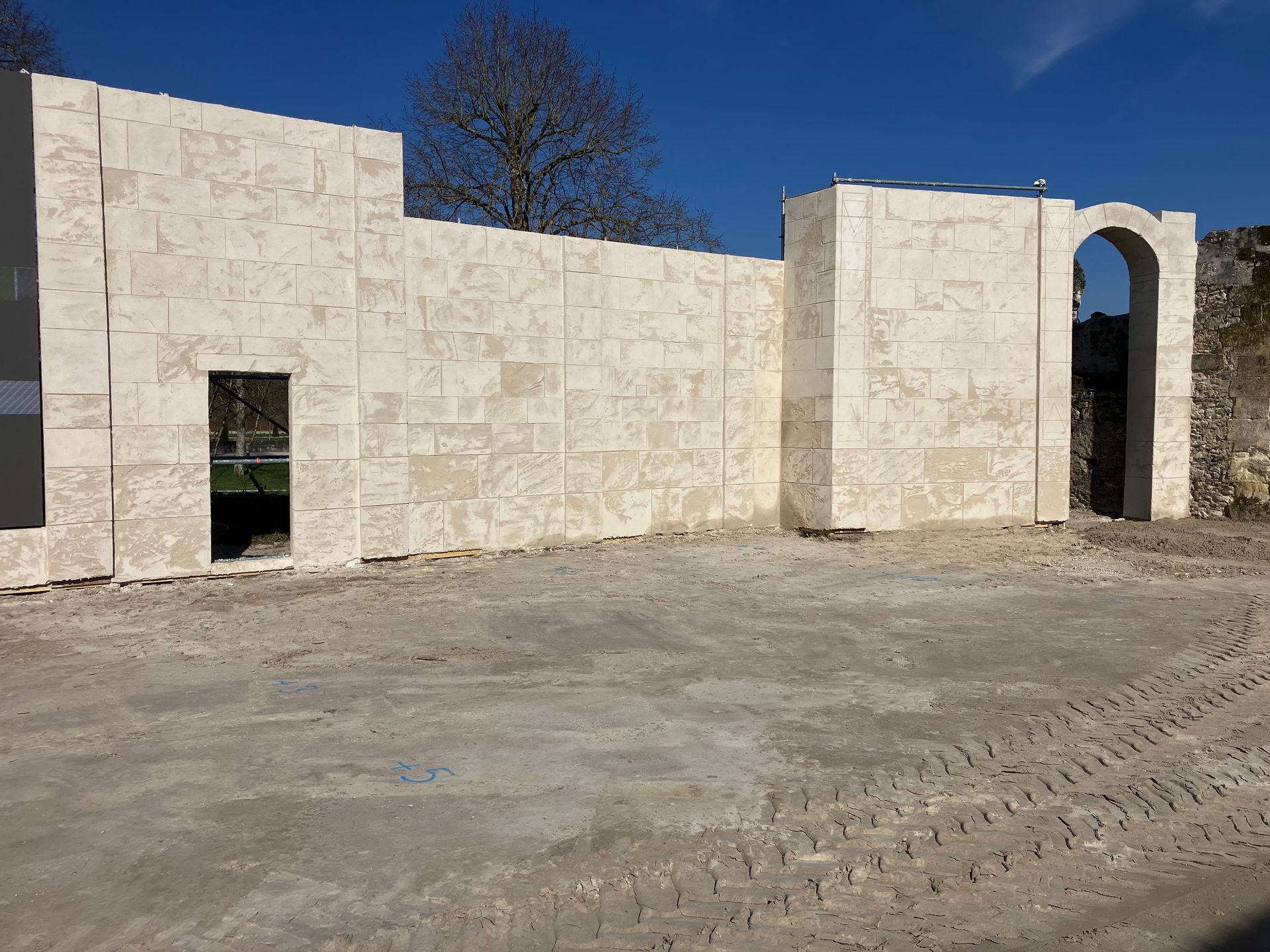 Édifice en pierre en construction, avec porte cintrée et murs inachevés, sur un terrain en terre battue.
