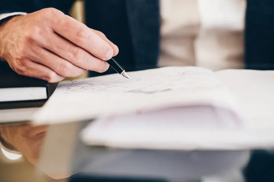 Firma manual de un documento en un escritorio con un bolígrafo, con papeles y un cuaderno borroso en primer plano.