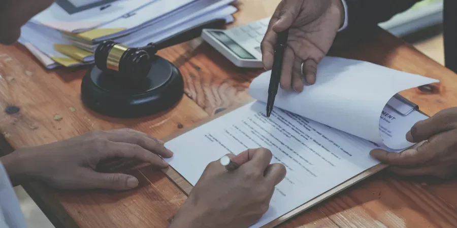 Manos firmando un documento en un escritorio con papeles, bolígrafo y sello.