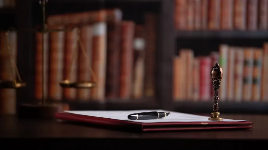 Pluma estilográfica sobre una carpeta roja encima de un escritorio, con estanterías borrosas al fondo.