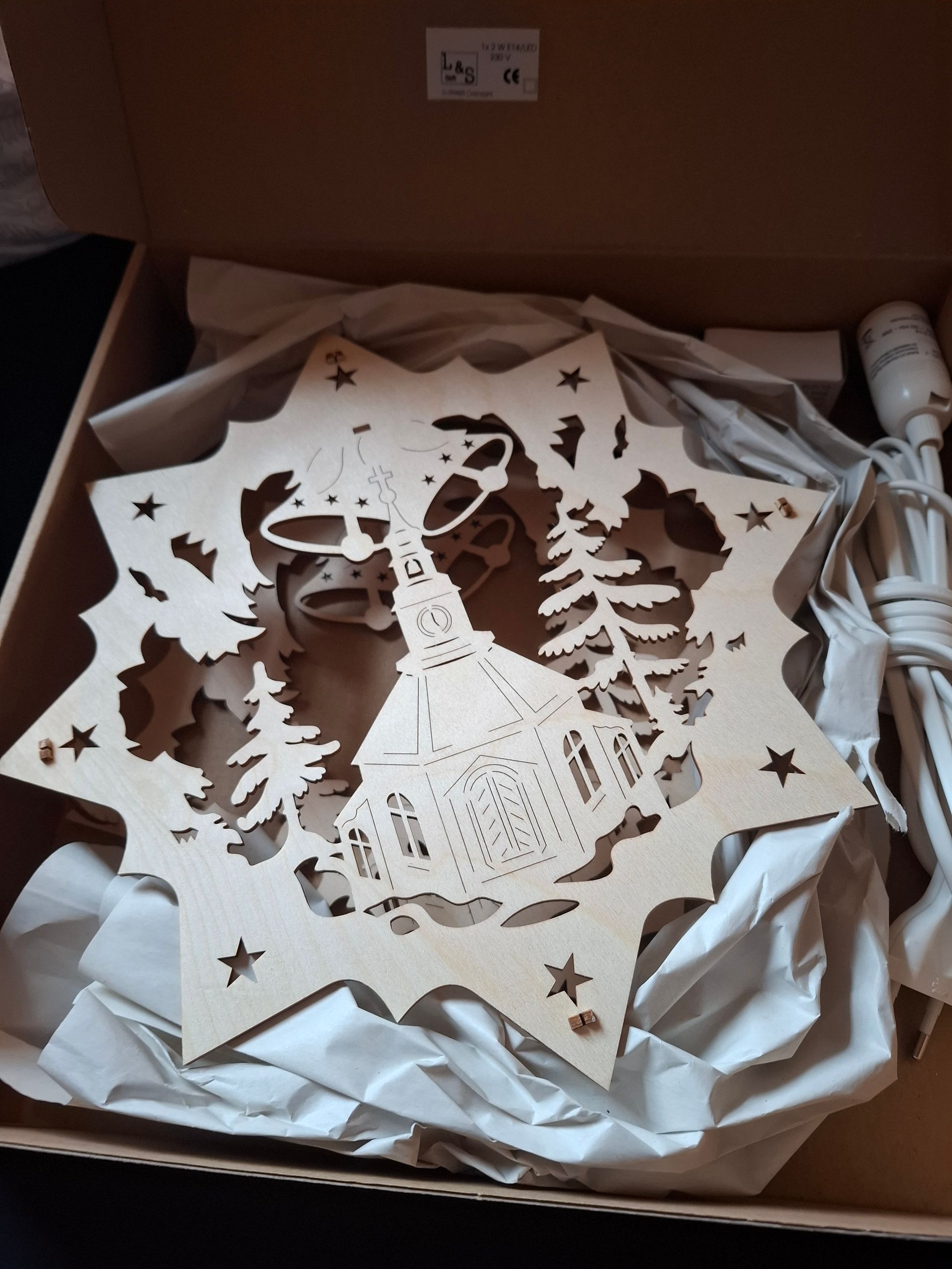 Weihnachtslicht aus Holz in Sternform mit ausgeschnittener Kirche, Bäumen und Sternen, in einem Karton mit zerknülltem Papier.
