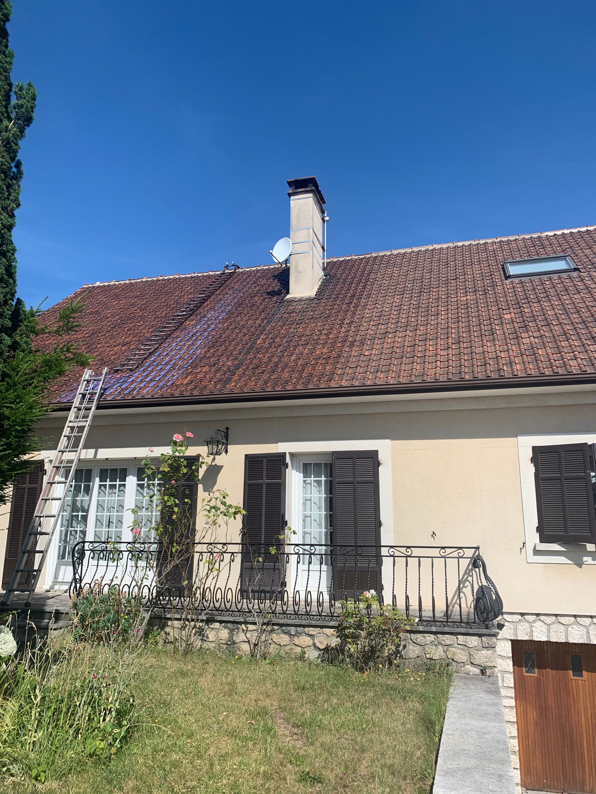 Petite maison au toit de tuiles rouges, aux volets blancs et à la cour avant envahie par la végétation, sous un ciel bleu.