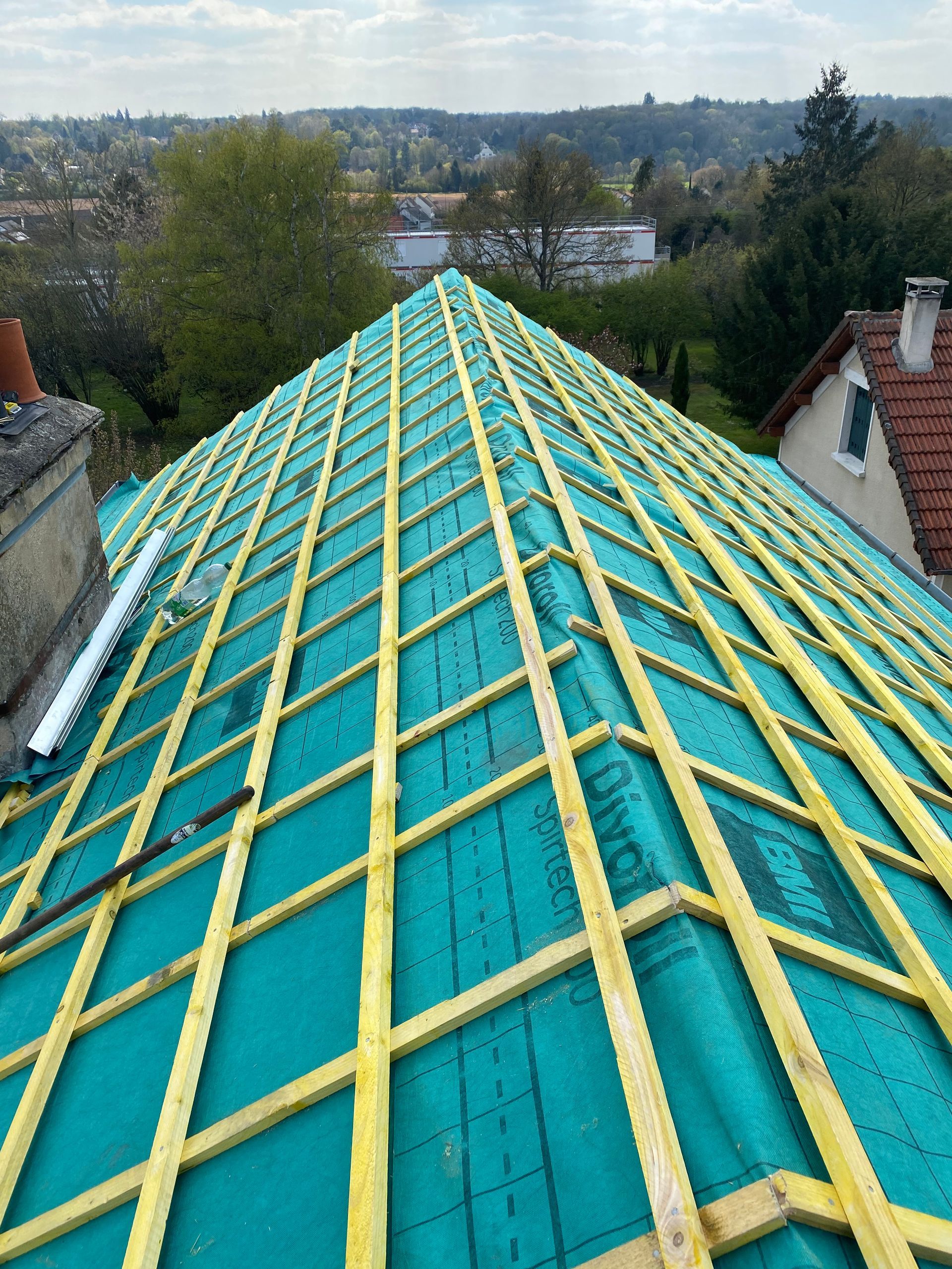 Lattes de toit jaunes au-dessus d'une toiture turquoise, vues depuis la crête entre les maisons surplombant les arbres et les maisons au loin.