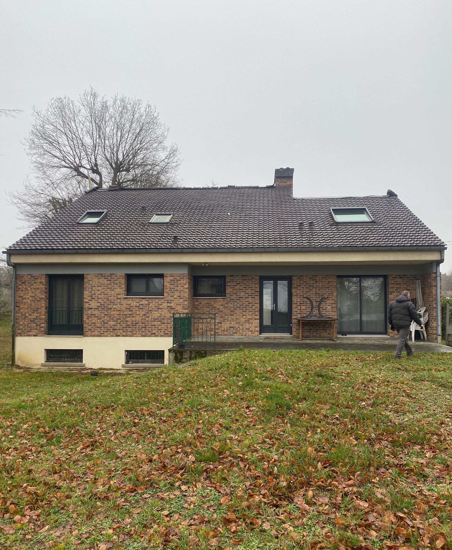 Maison en briques avec un toit de tuiles sombres et une pelouse recouverte de feuilles sous un ciel couvert