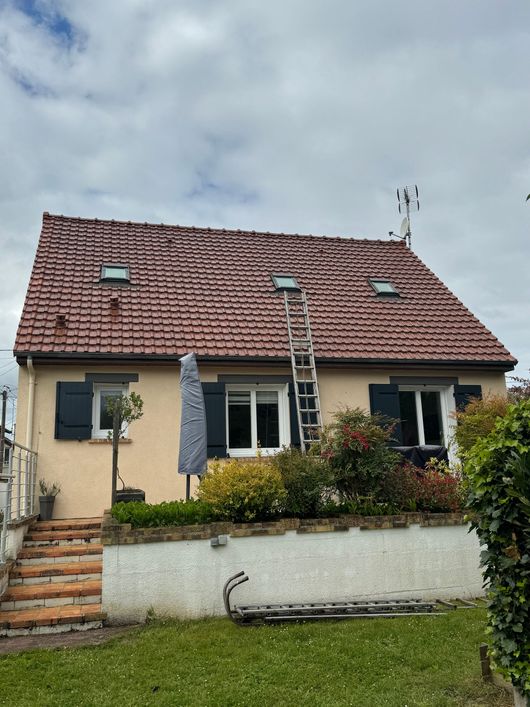 Petite maison couleur crème avec toit de tuiles rouges, jardin à l'avant, parasol sur la terrasse et marches en pierre sous un ciel nuageux.