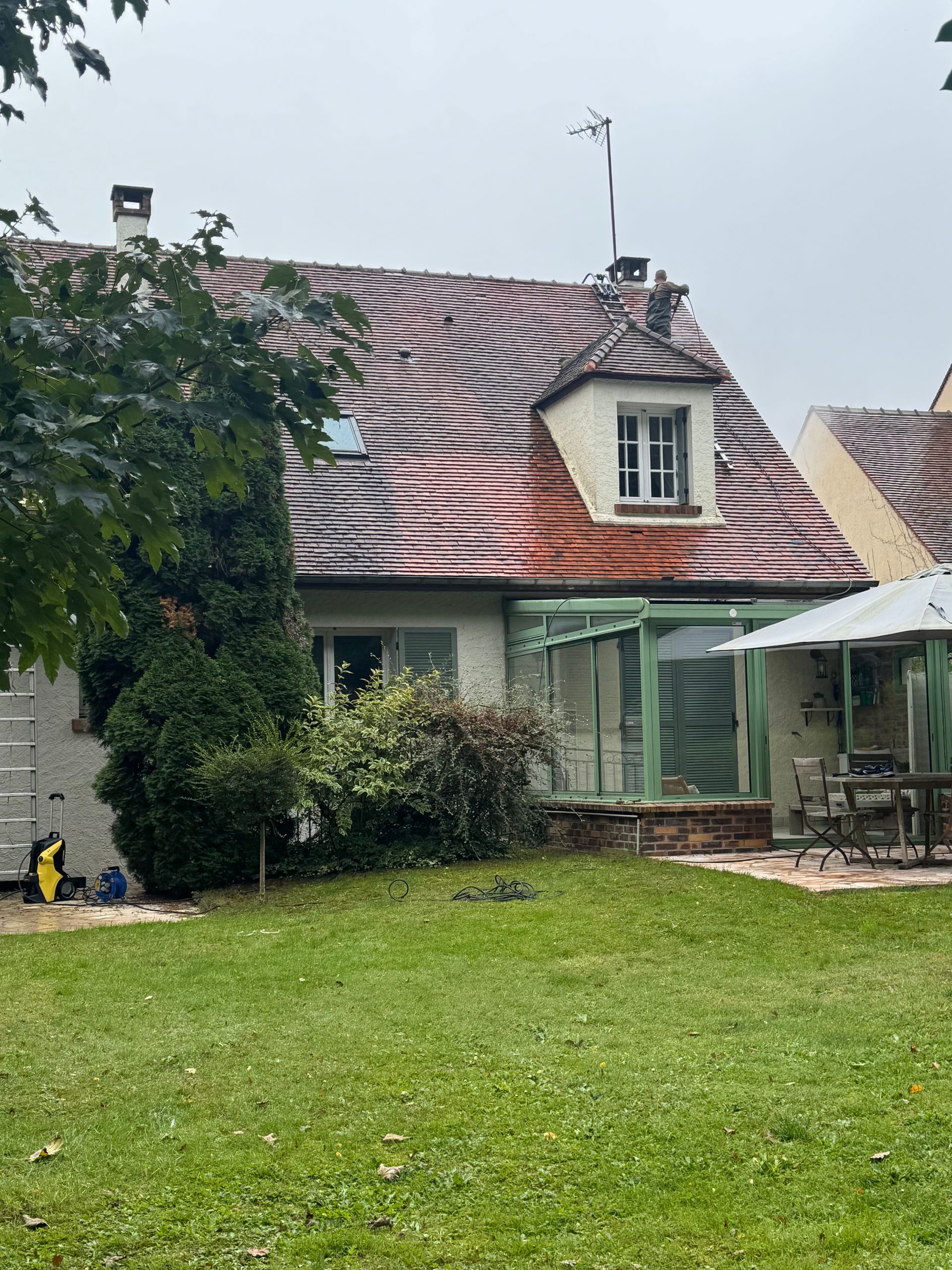Maison au toit de tuiles rouges, aux volets verts et au patio donnant sur une pelouse sous un ciel couvert.