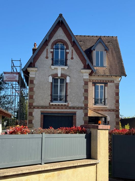 Maison en pierre de trois étages avec toit rouge, clôture grise et parterres de fleurs sous un ciel bleu clair