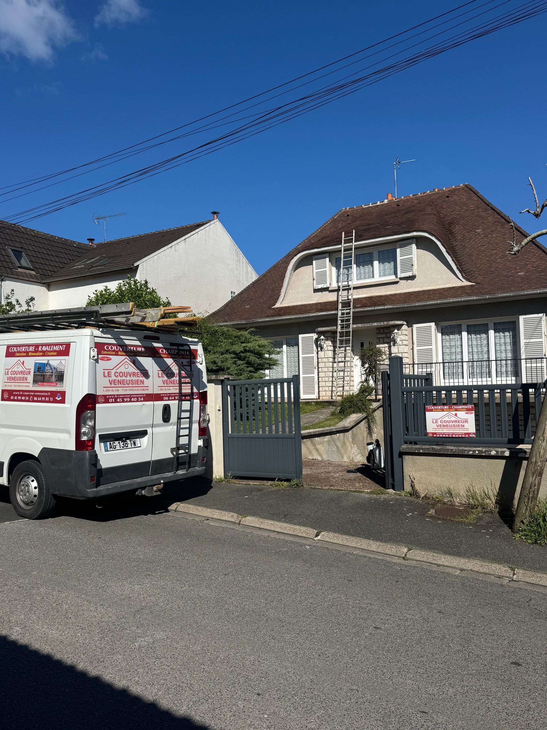 Une camionnette blanche est garée devant une maison au toit de tuiles rouges et au portail ouvert, dans une rue ensoleillée.