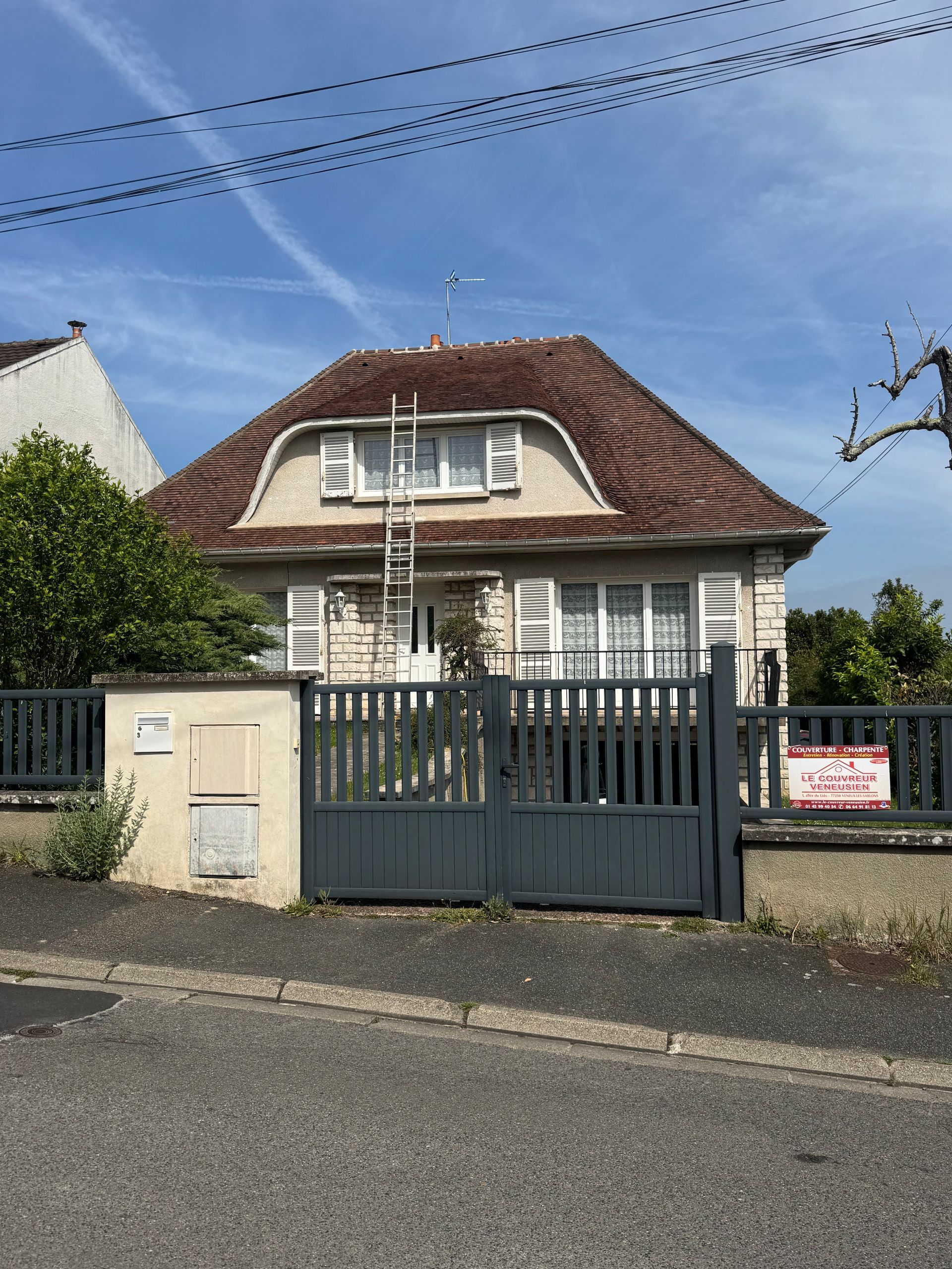 Maison blanche au toit rouge derrière un portail et une clôture gris, dans une rue ensoleillée