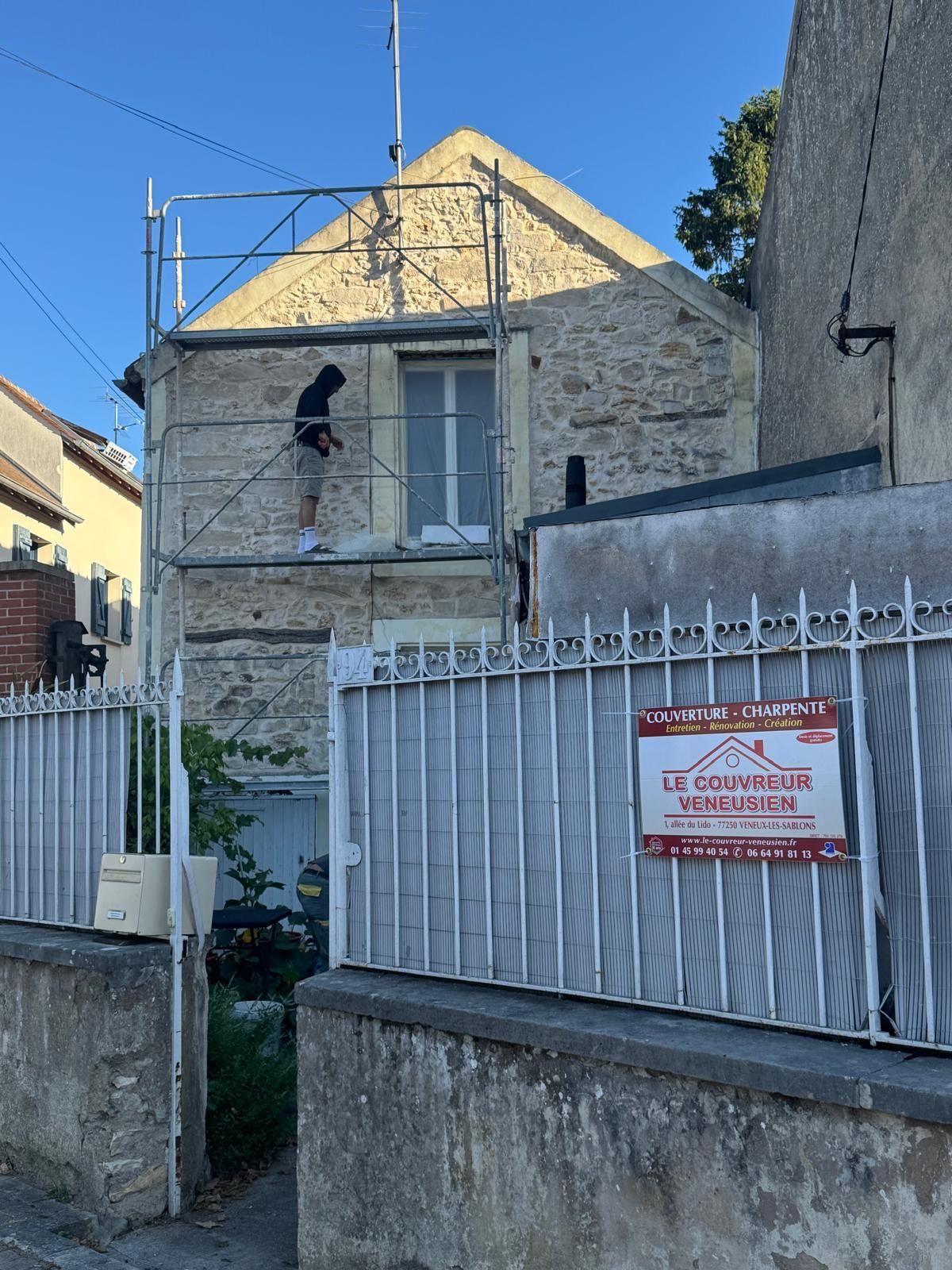 Maison en pierre avec un ouvrier sur un échafaudage derrière une clôture métallique et un panneau d'avertissement