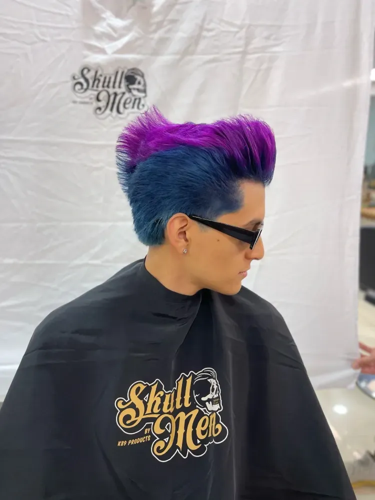 Un hombre con cabello azul y morado lleva gafas de sol y una capa.