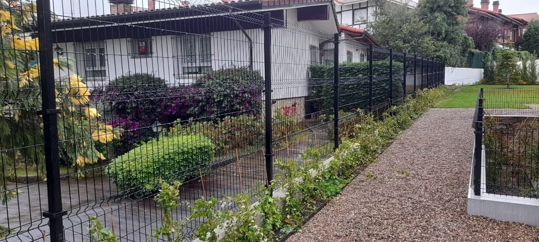 Una valla rodea un exuberante jardín verde con una casa al fondo.