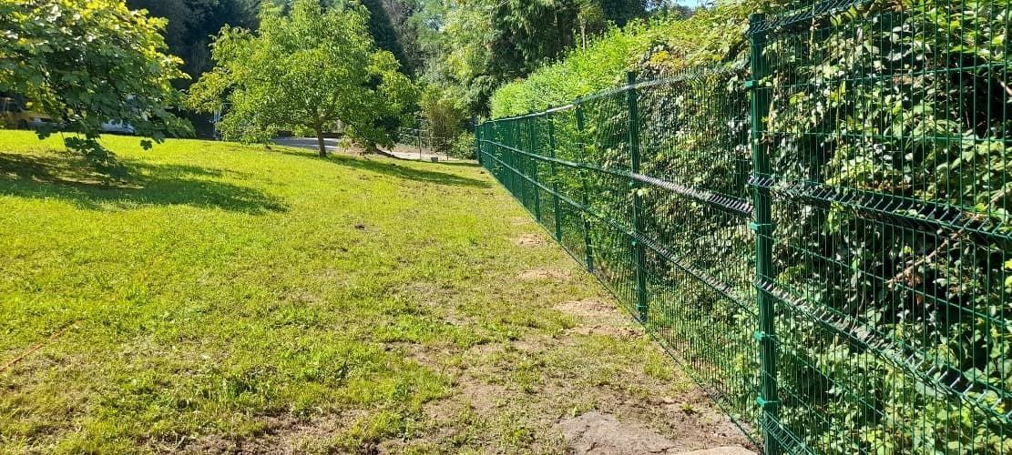 Una valla verde rodea un campo de hierba con árboles al fondo.