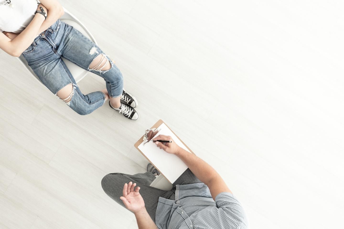Una persona con jeans rotos se sienta frente a alguien que escribe en un portapapeles; habitación blanca y luminosa.