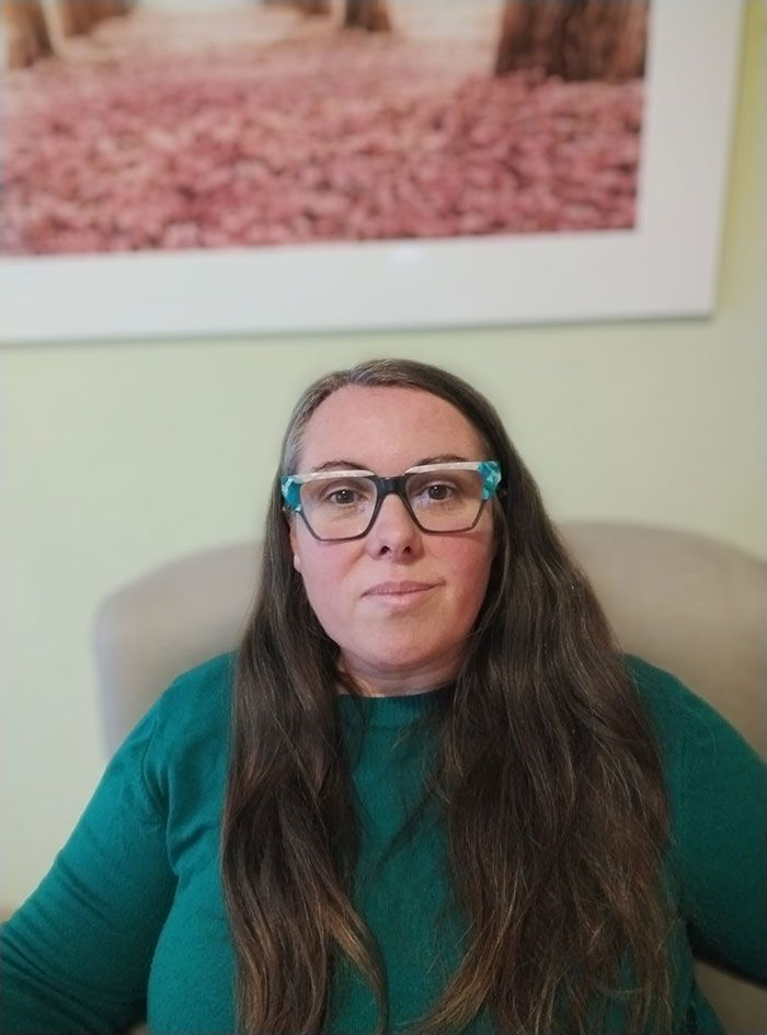 Mujer con cabello largo castaño, con gafas y un suéter verde azulado, sentada en una silla, con una foto detrás de ella.