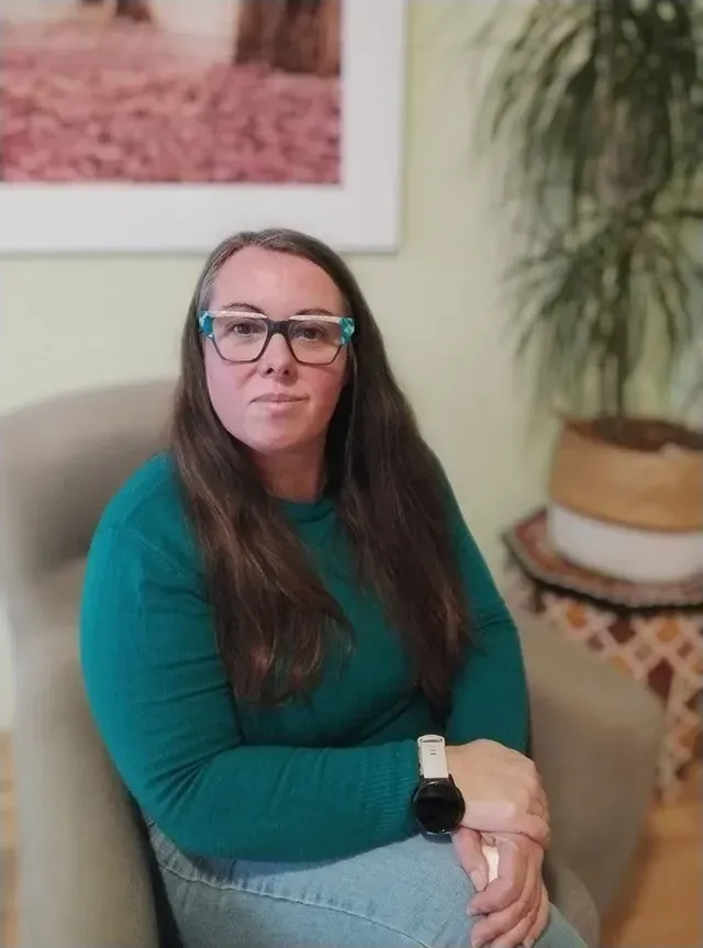 Mujer sentada en una silla en el interior, con gafas y una blusa color turquesa, con una planta en maceta y una obra de arte detrás de ella.