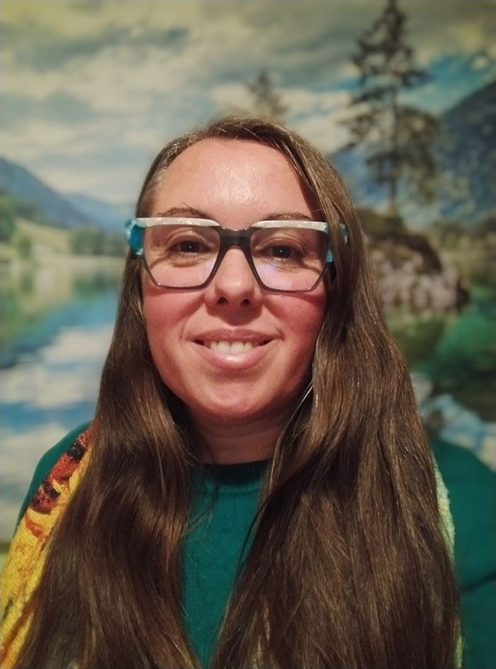 Mujer con gafas, sonriente, con cabello castaño largo y camisa verde. Montañas pintadas al fondo.