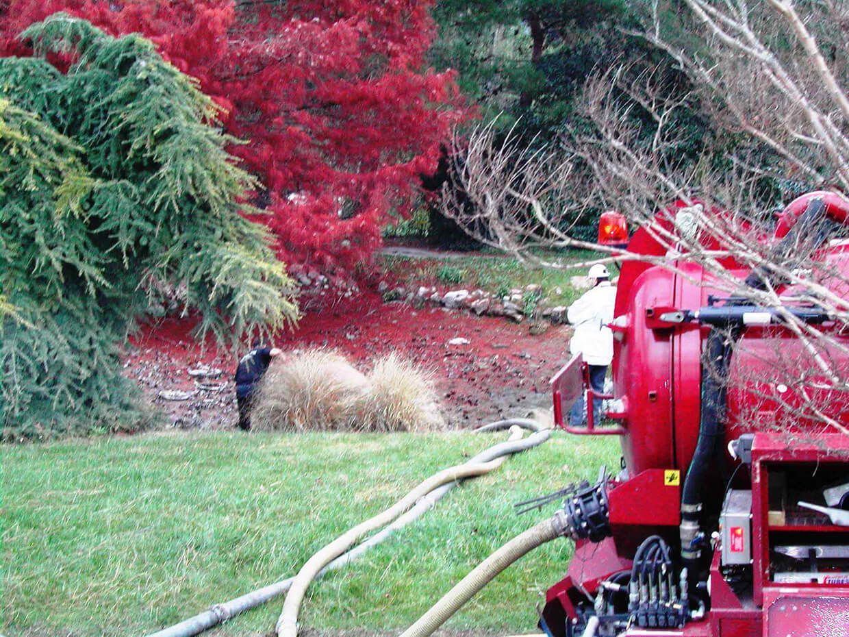 L'arrière d'un camion d'assainissement rouge projetant des résidus de tonte à côté d'une pelouse verte et d'arbustes rouges d'automne.
