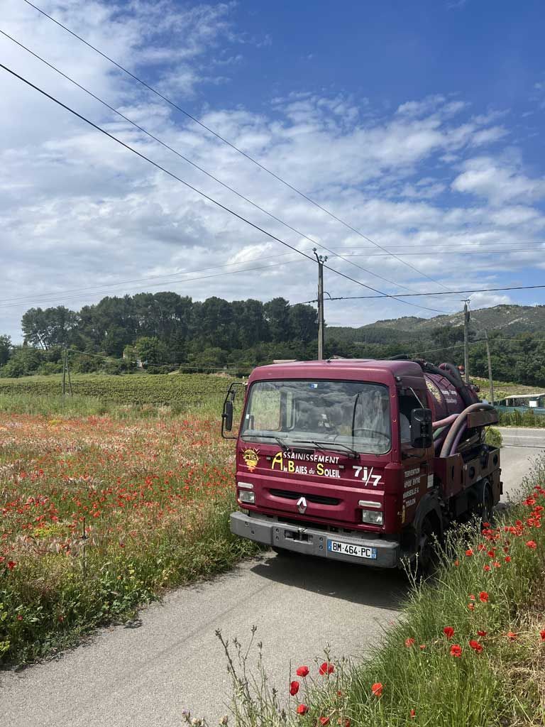 Camion d'assainissement bordeaux sur une route de campagne étroite, à côté d'un champ de fleurs sauvages sous un ciel nuageux