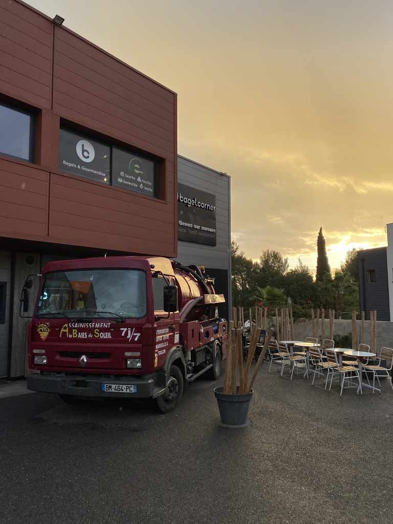 Un camion d'assainissement rouge est stationné devant un immeuble au coucher du soleil, avec une terrasse de café à proximité.