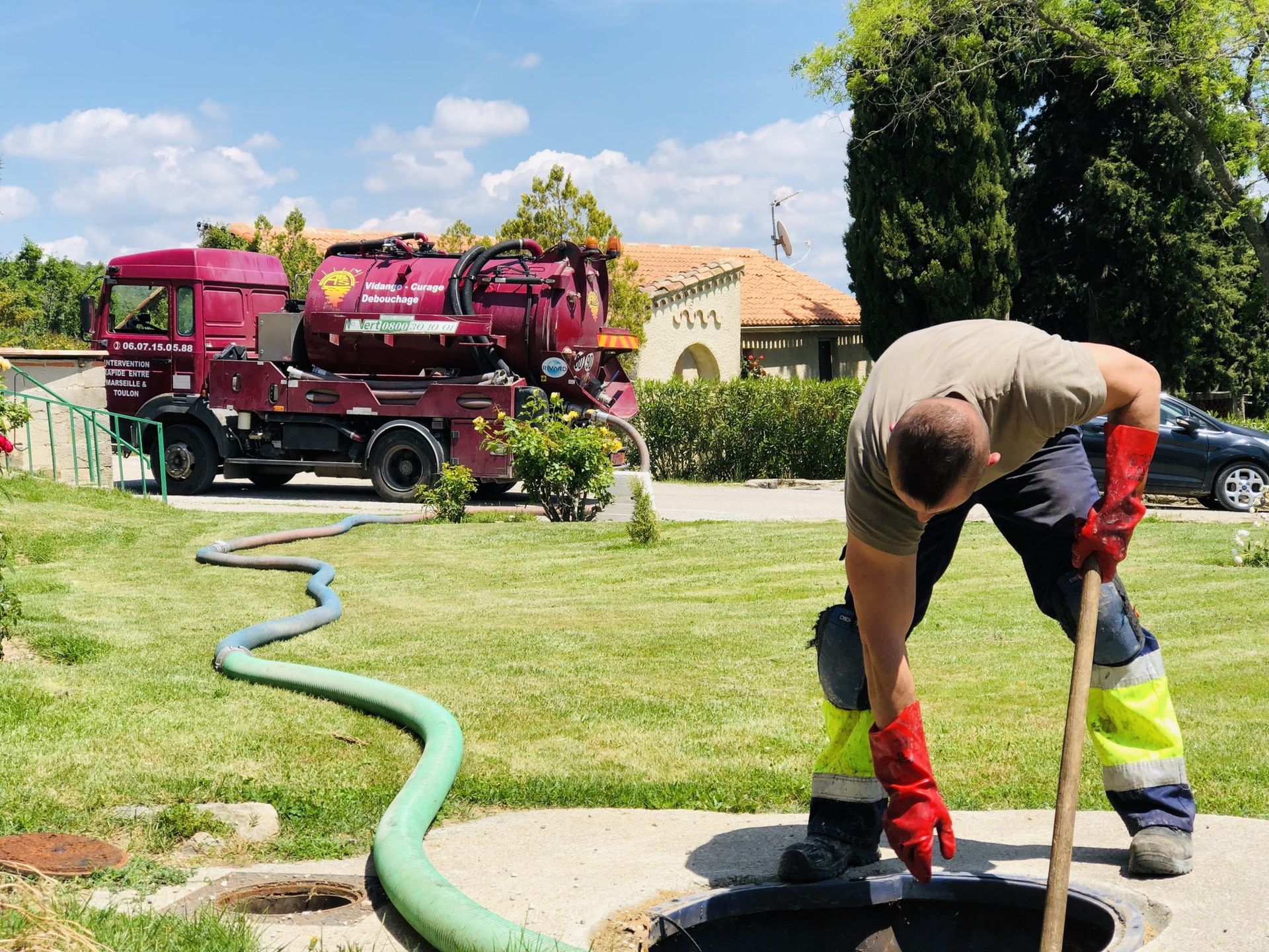 Un ouvrier utilise un enrouleur de tuyau d'arrosage dans une cour herbeuse, à côté d'un camion d'assainissement rouge et d'une maison.