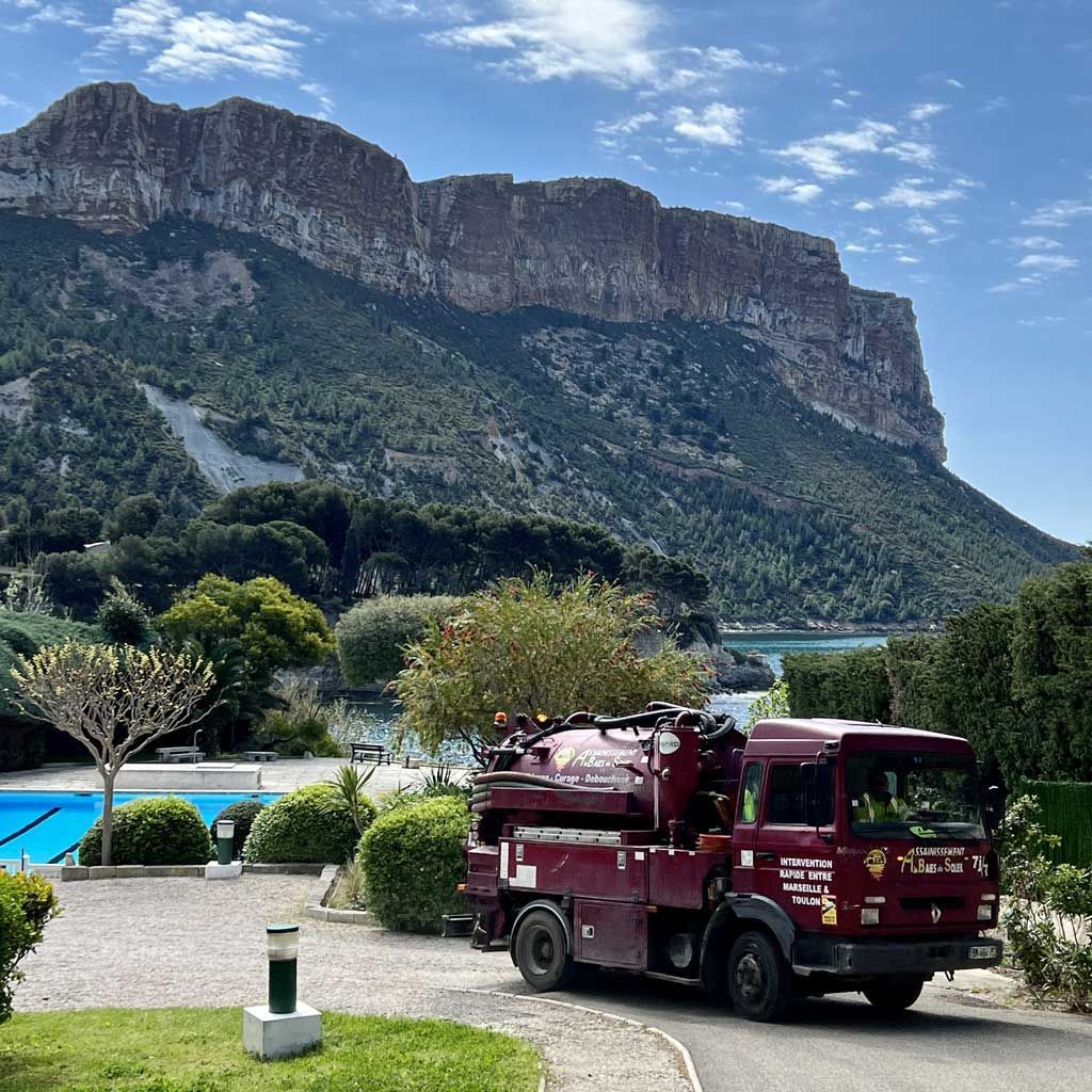 Camionnette d'assainissement bordeaux garée au bord d'une piscine, avec une falaise montagneuse en arrière-plan.