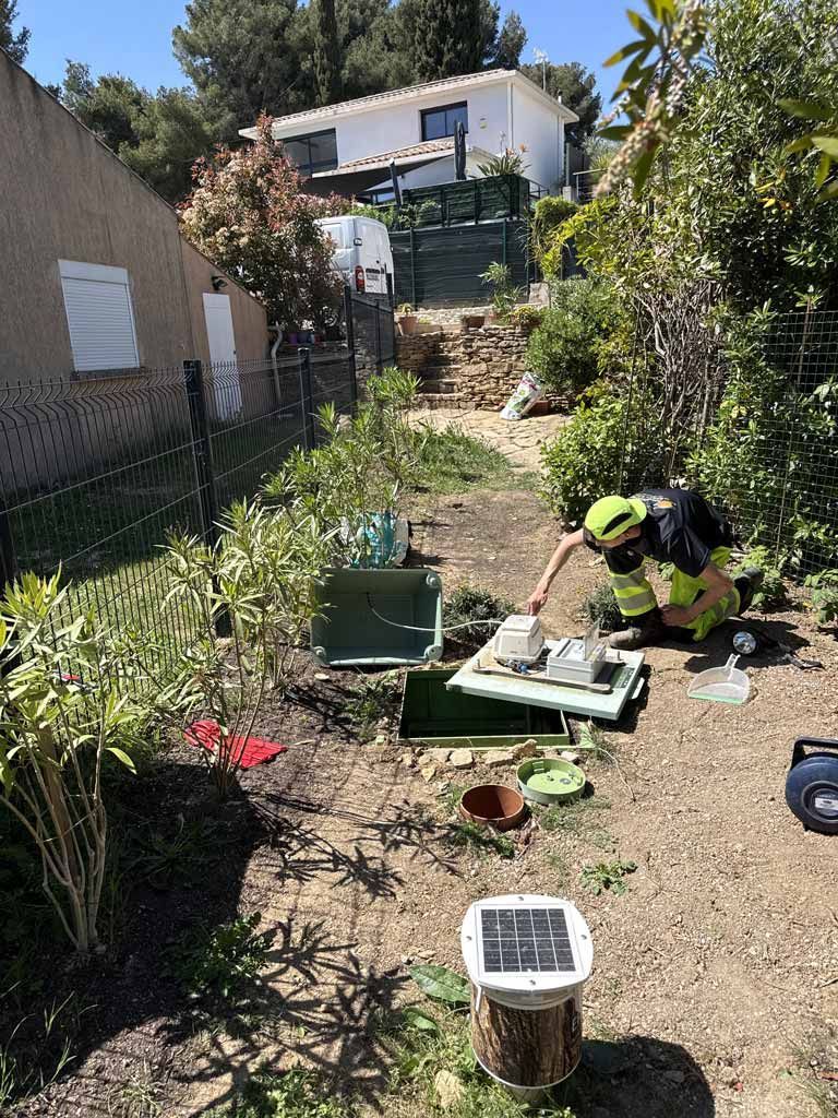 Un ouvrier agenouillé dans un jardin, près d'outils et de pavés, avec une maison et des plantes en arrière-plan.