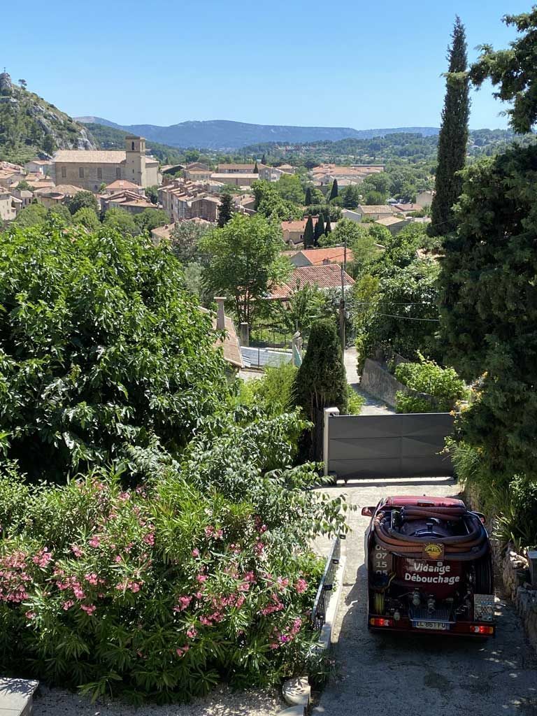 Vue sur un village à flanc de colline avec des arbres, des bâtiments aux toits rouges et un petit véhicule sur une route au premier plan.