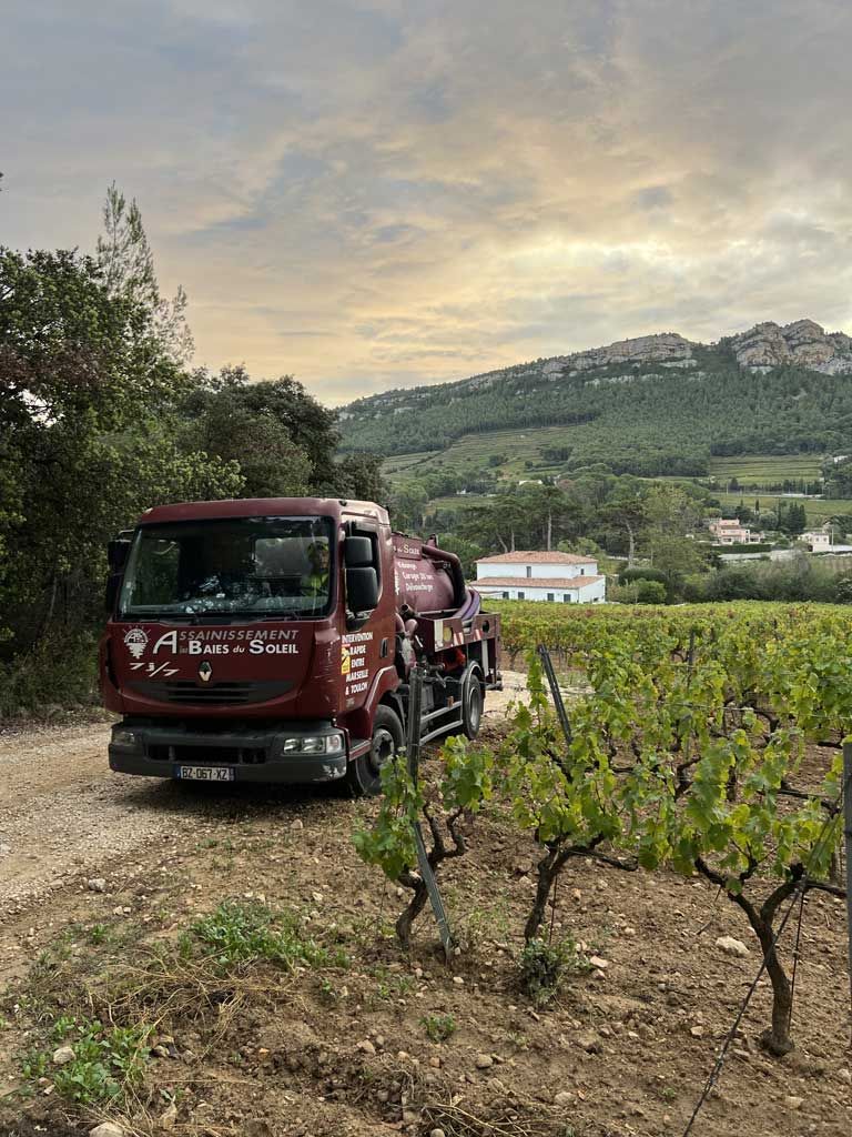 Un camion bordeaux d'assainissement est garé sur un chemin de terre bordé de vignes, avec des collines verdoyantes et un ciel nuageux en arrière-plan.